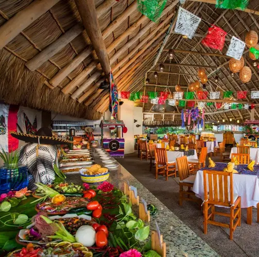 Restaurante mexicano con techo de palma, mesas decoradas y buffet con comida típica. Banderas de papel adornan el techo.
