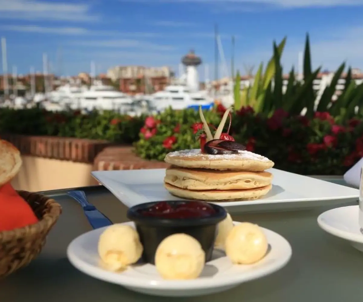 Pancakes servidos con mantequilla y mermelada en una terraza con vista al puerto deportivo y barcos al fondo.