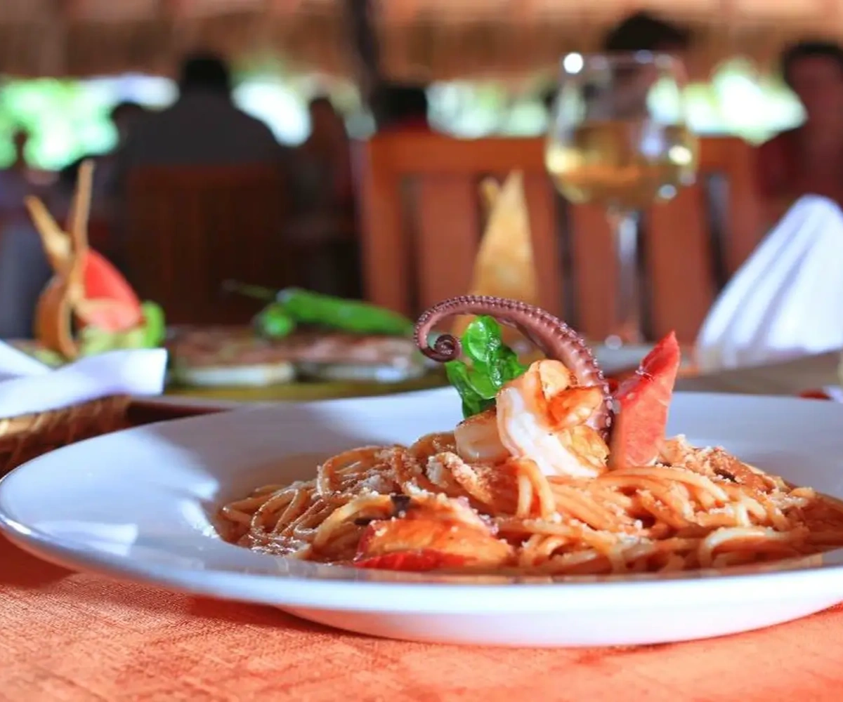 Plato de pasta con mariscos y pulpo, servido en un restaurante, con una copa de vino al fondo.
