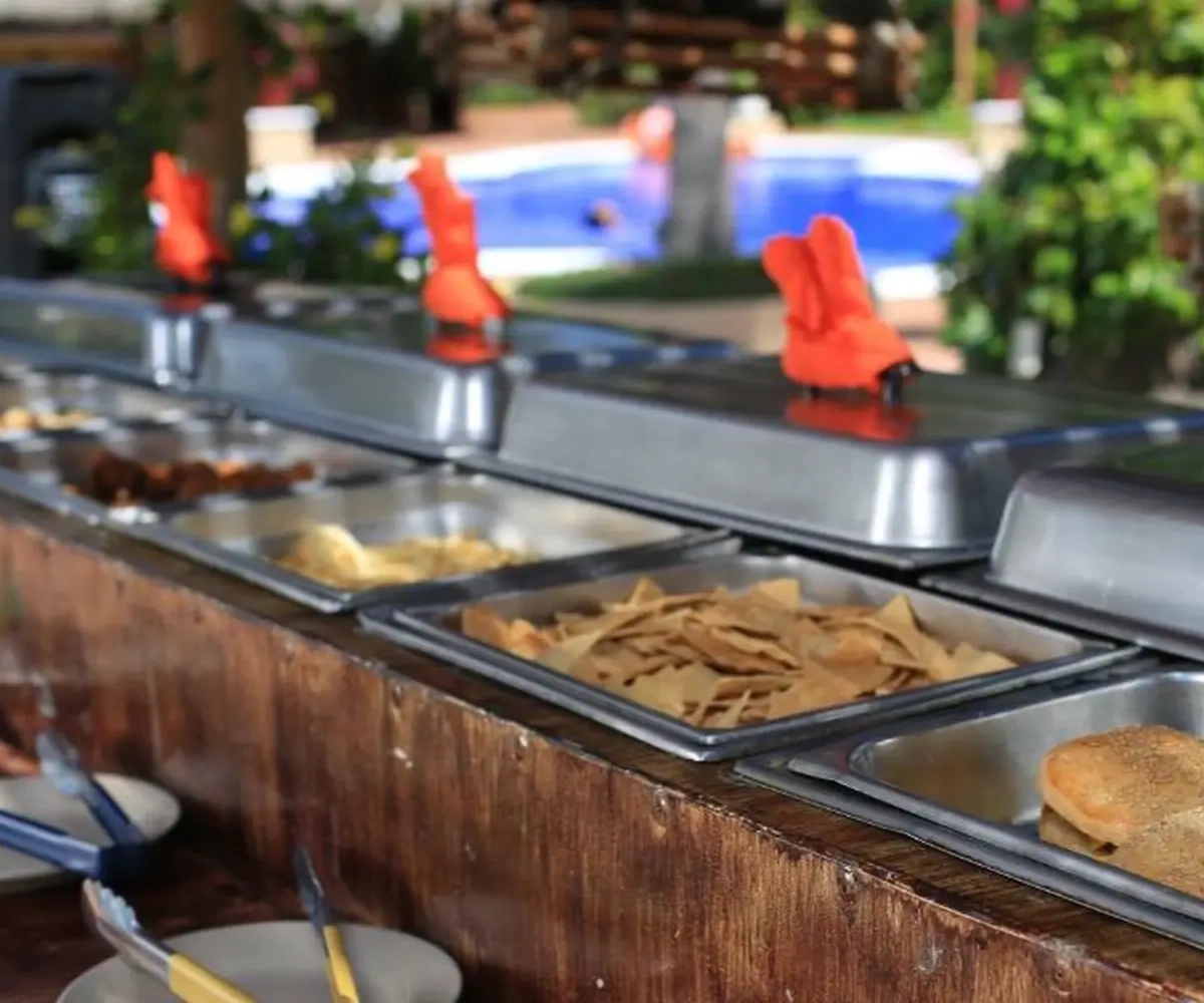 Buffet al aire libre con bandejas de comida diversa y pinzas, con fondo de piscina y vegetación.