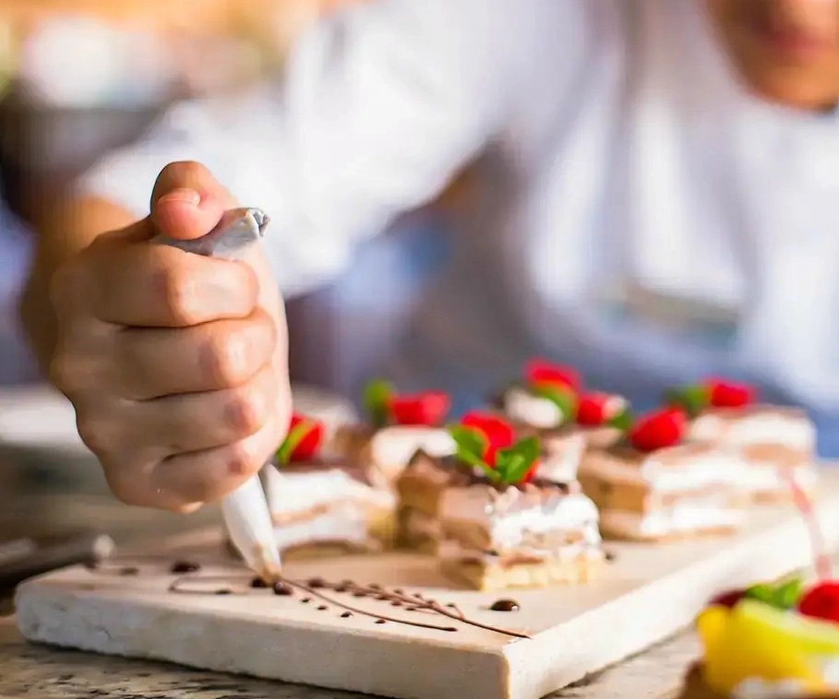 Mano decorando postres con una manga pastelera sobre una tabla blanca, con porciones y fresas de fondo.