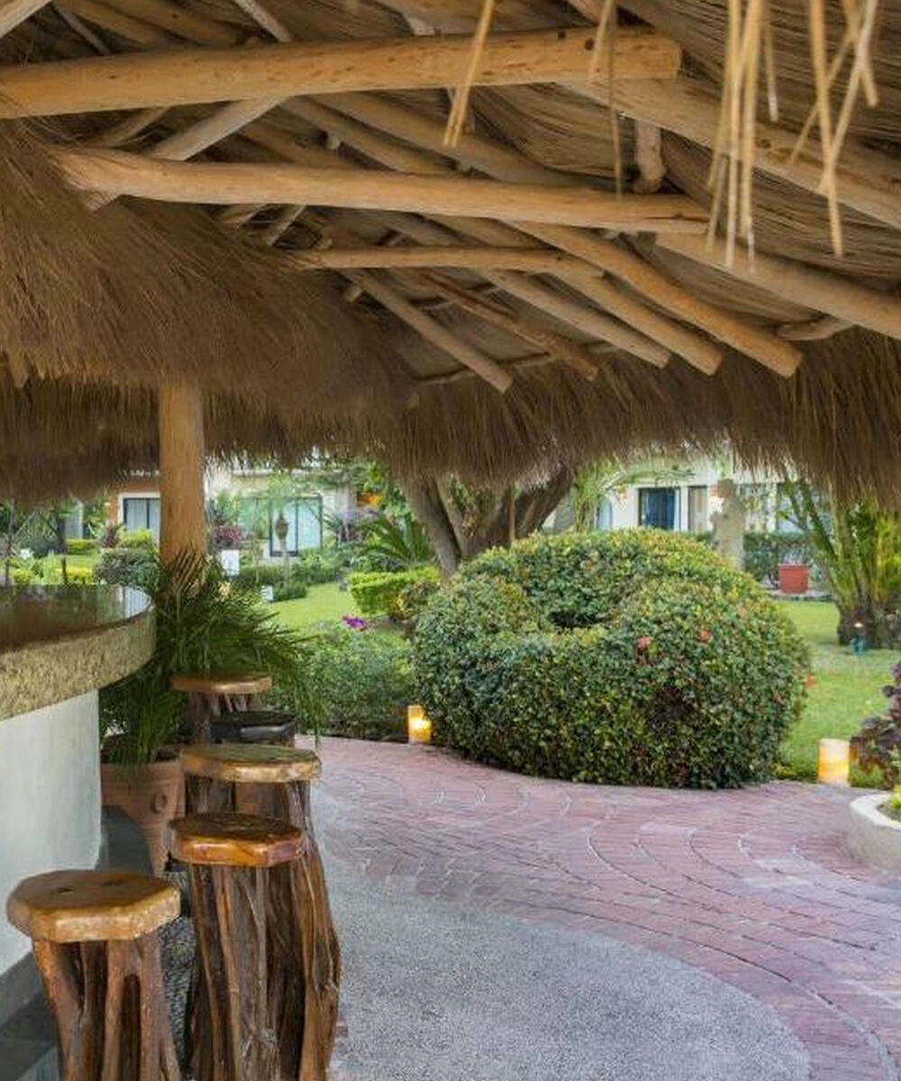 Barra al aire libre con techo de palapa y taburetes de madera, rodeada de jardines y caminos de piedra.
