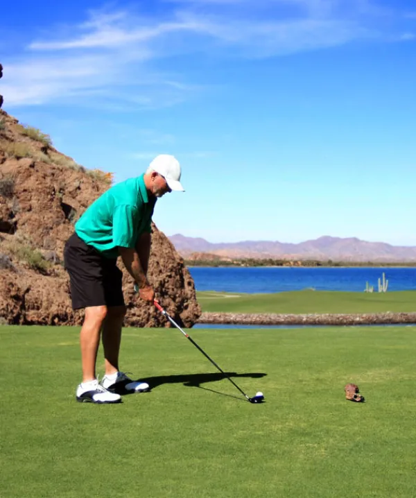 Hombre jugando golf en un campo cercano a un lago y monta&ntilde;as, con cielo despejado.