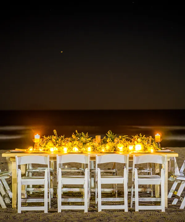 Mesa decorada en la playa de noche, iluminada con velas y flores.