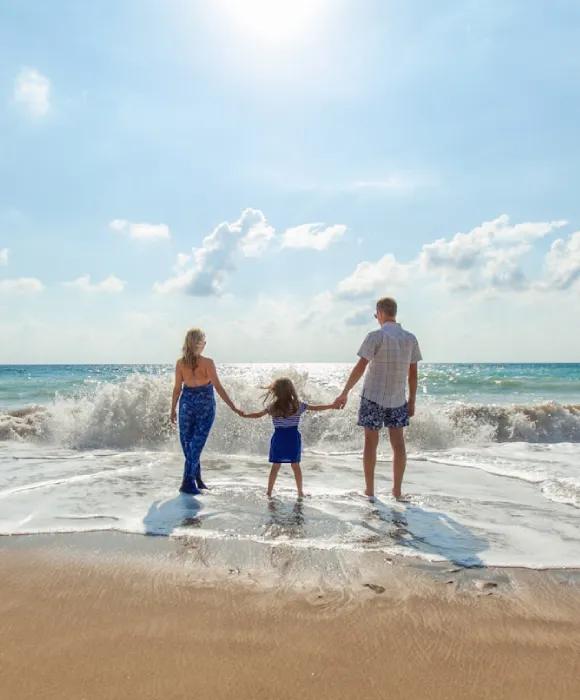 Familia de tres personas disfruta de la playa, cogidos de la mano frente al mar bajo el sol.
