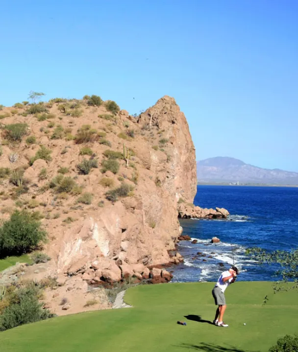 Persona jugando golf junto a un acantilado con vista al mar y monta&ntilde;as en el fondo.
