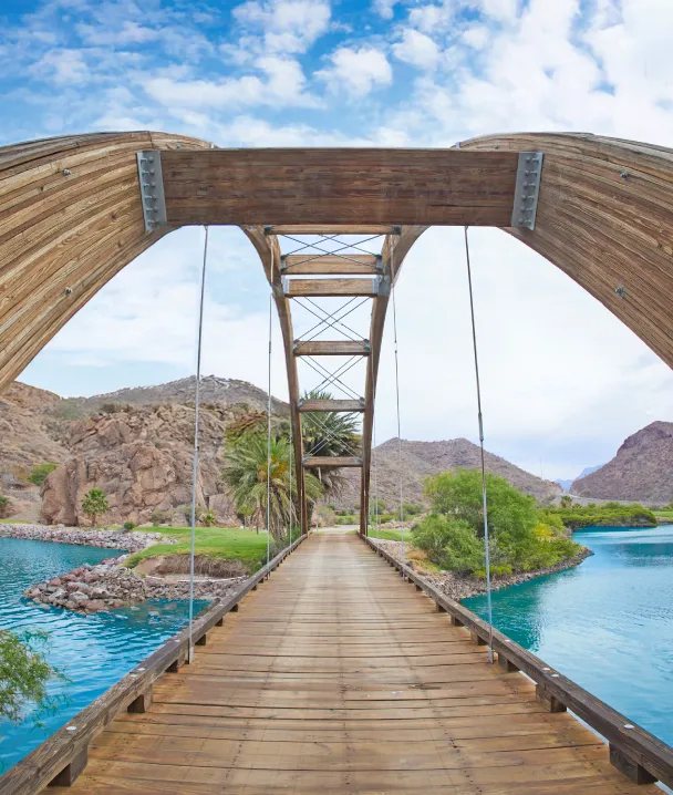 Puente de madera sobre agua azul, rodeado de monta&ntilde;as y vegetaci&oacute;n bajo un cielo parcialmente nublado.