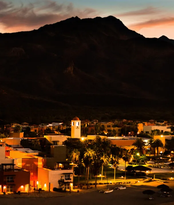 Pueblo iluminado al anochecer, con monta&ntilde;as oscuras de fondo y cielo con tonos c&aacute;lidos.