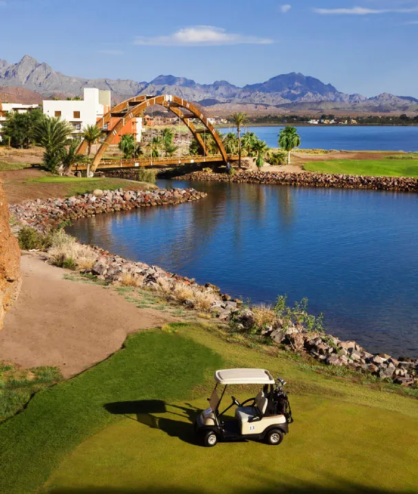 Paisaje de campo de golf con lago, puente de madera y carrito en primer plano. Monta&ntilde;as al fondo.