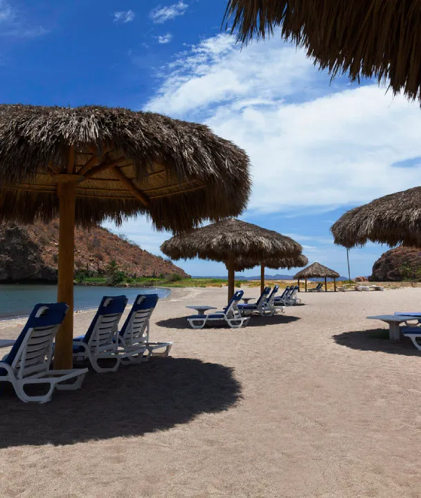 Sombrillas de paja y tumbonas en una playa de arena bajo un cielo azul con monta&ntilde;as al fondo.