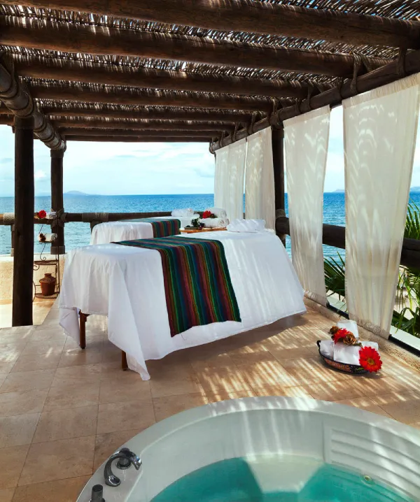 Pareja de camillas de masaje bajo p&eacute;rgola junto al mar, con jacuzzi al frente.