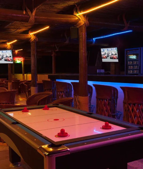 Sala de juegos con mesa de hockey de aire, barra iluminada y pantallas con autos de carreras.