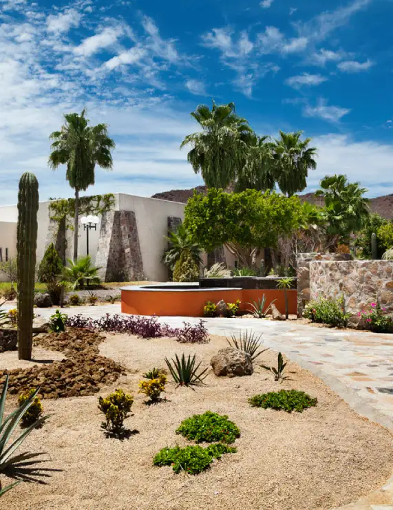 Jard&iacute;n des&eacute;rtico con cactus, palmeras y camino frente a edificio de piedra con cielo azul.