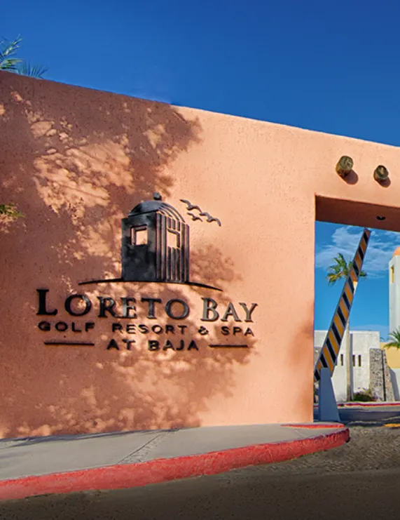 Entrada de Loreto Bay Golf Resort & Spa, muro color salm&oacute;n con el logo y cielo azul.