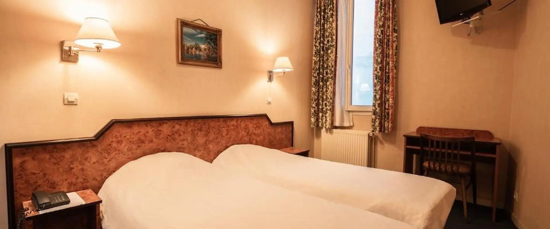 Chambre double avec lits jumeaux, lampe murale, tableau, bureau, télévision et fenêtre.