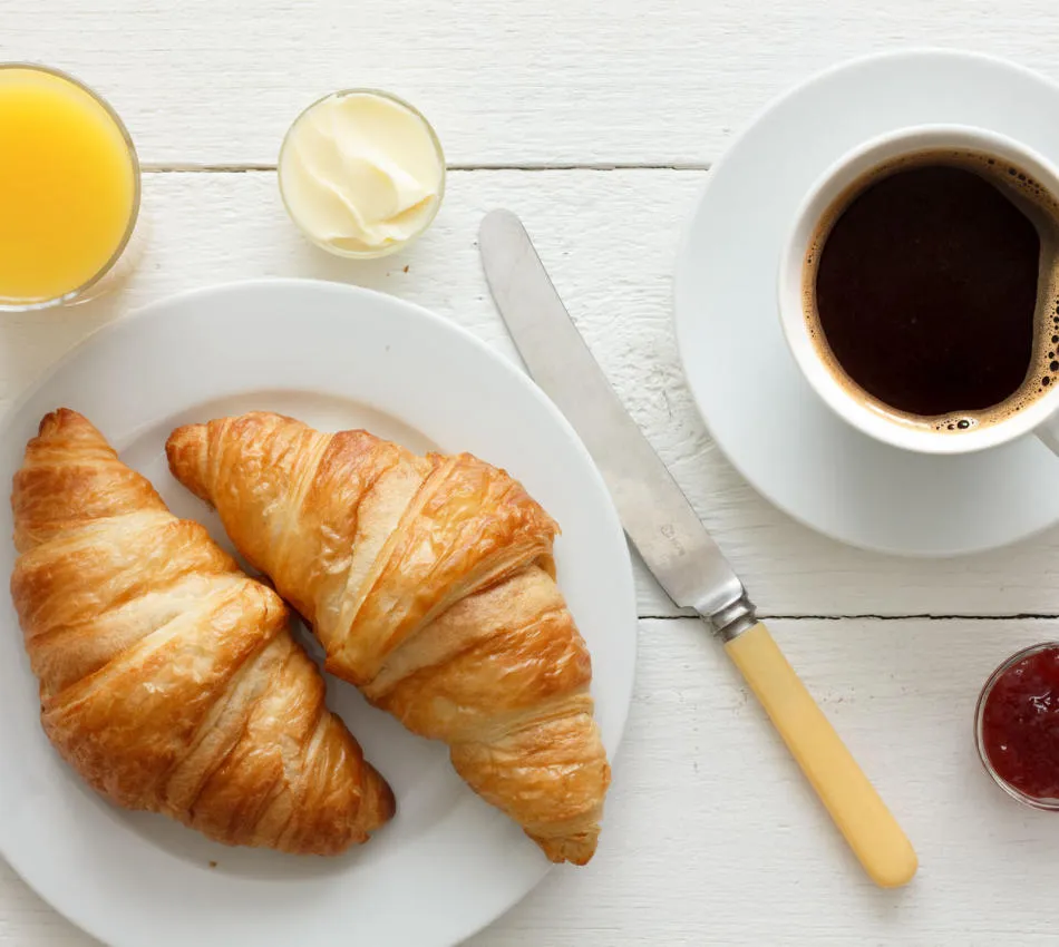 Petit déjeuner avec croissants, café, jus d’orange, beurre et confiture sur table blanche.