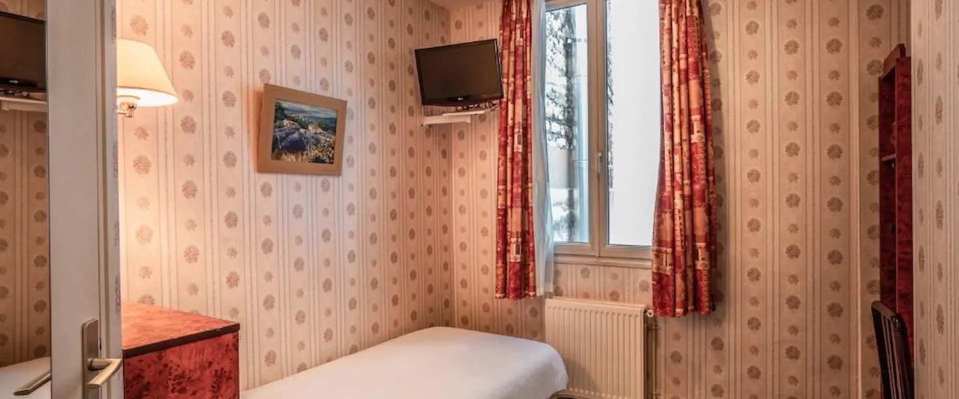 Petite chambre avec lit simple, tapisserie vintage, tableau, téléviseur et fenêtre.