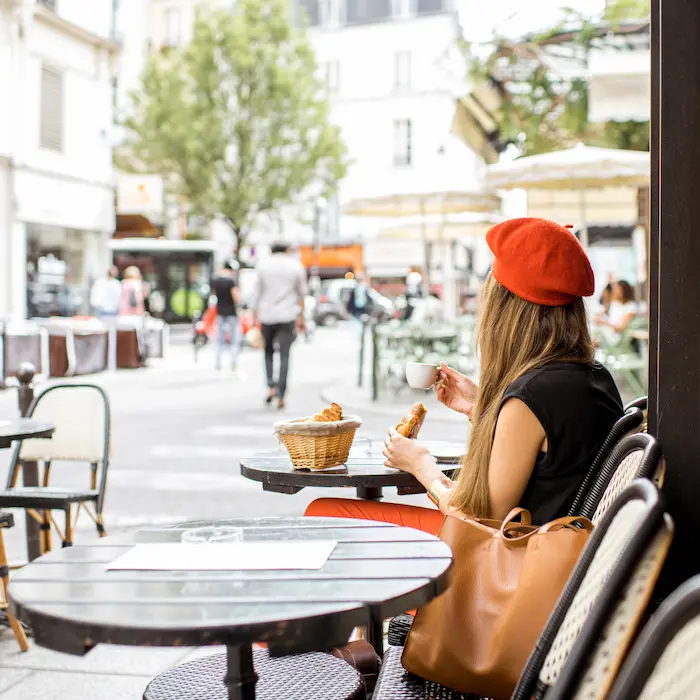 Femme buvant un café en terrasse, portant un béret rouge et mangeant un croissant.