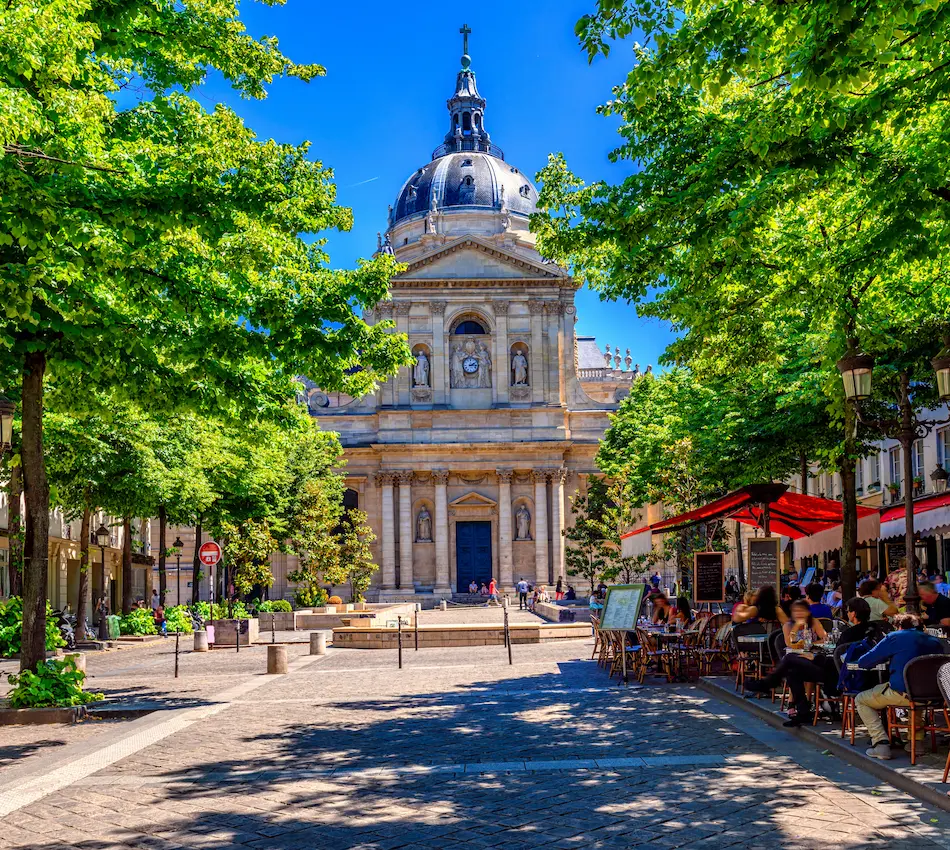 Place animée avec terrasse de café et bâtiment ancien sous un ciel bleu.