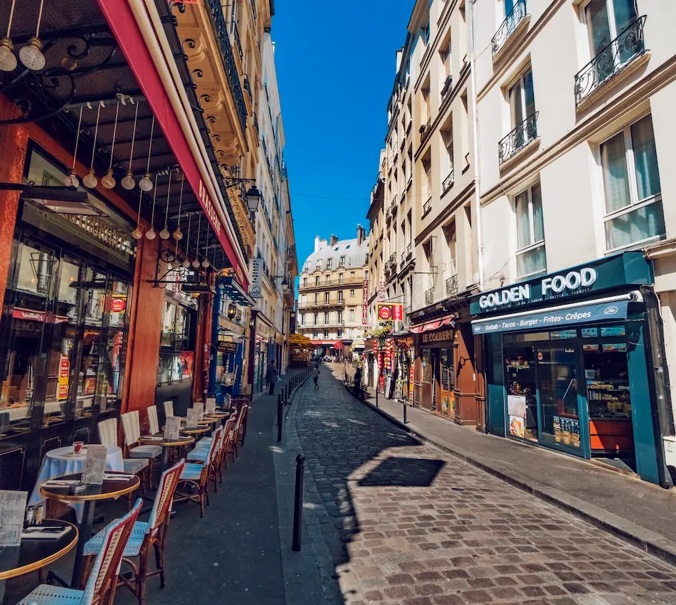 Rue pavée animée avec cafés et restaurants, ciel bleu.