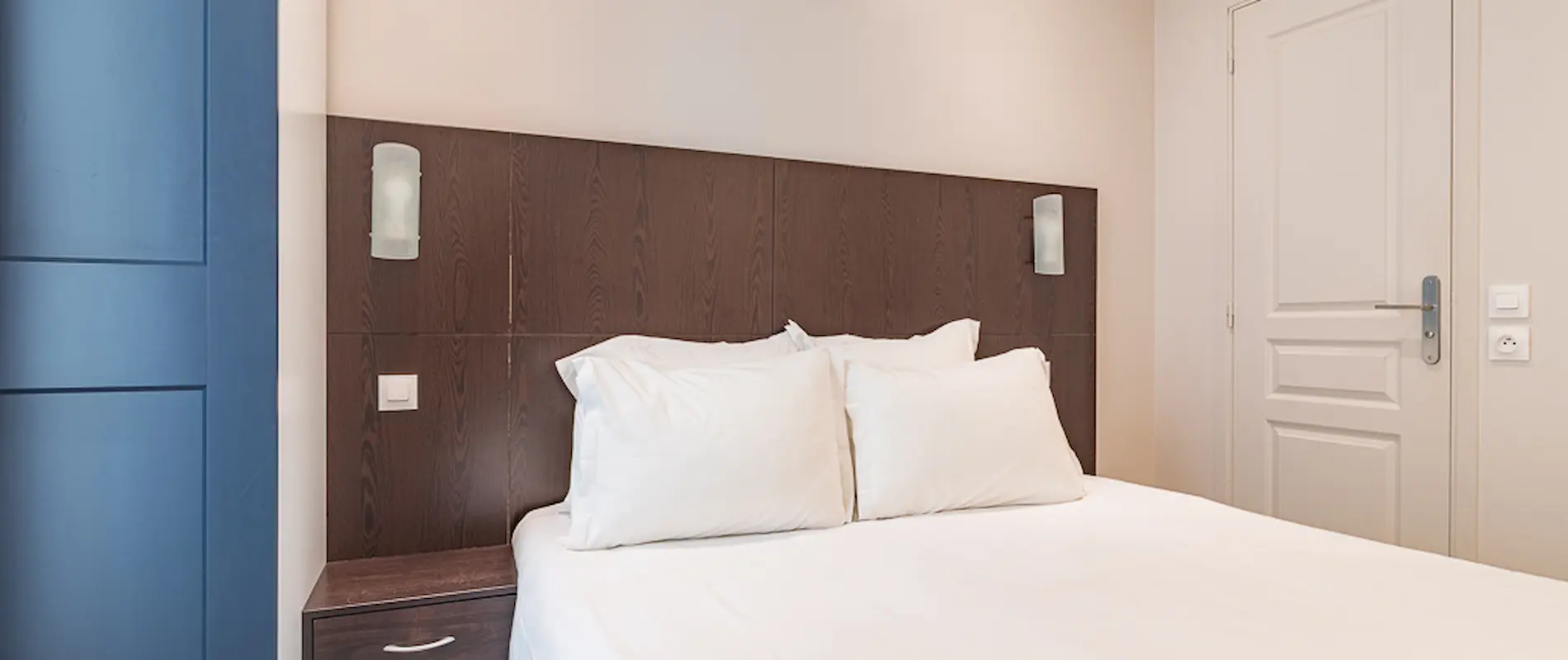 Lit double avec tête de lit en bois, deux lampes, et mur bleu à gauche.