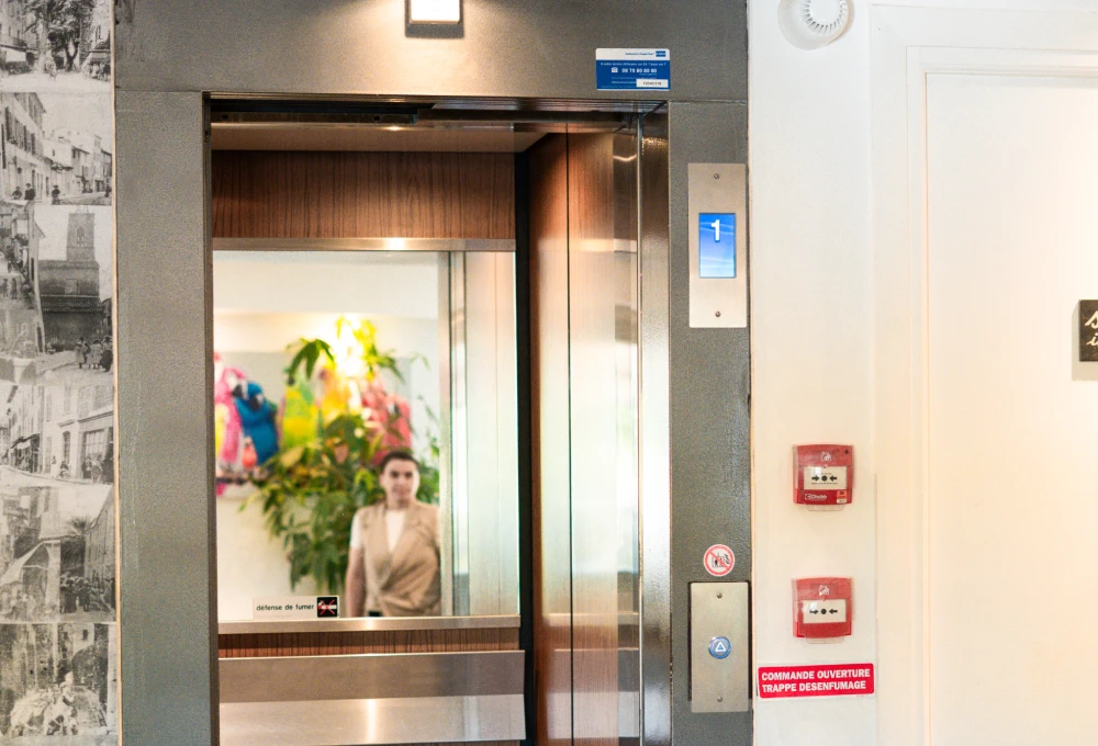 Un ascenseur ouvert reflète une personne floue; des plantes et couleurs vives en arrière-plan.