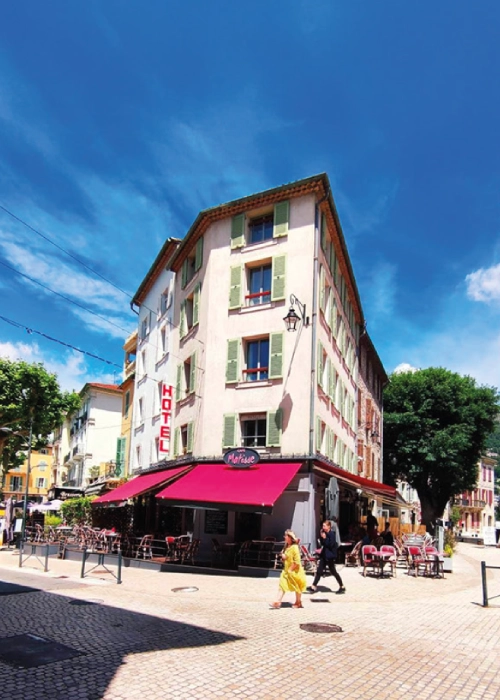 Immeuble d'angle avec hôtel et café, ciel bleu, personnes marchant sur rue pavée.