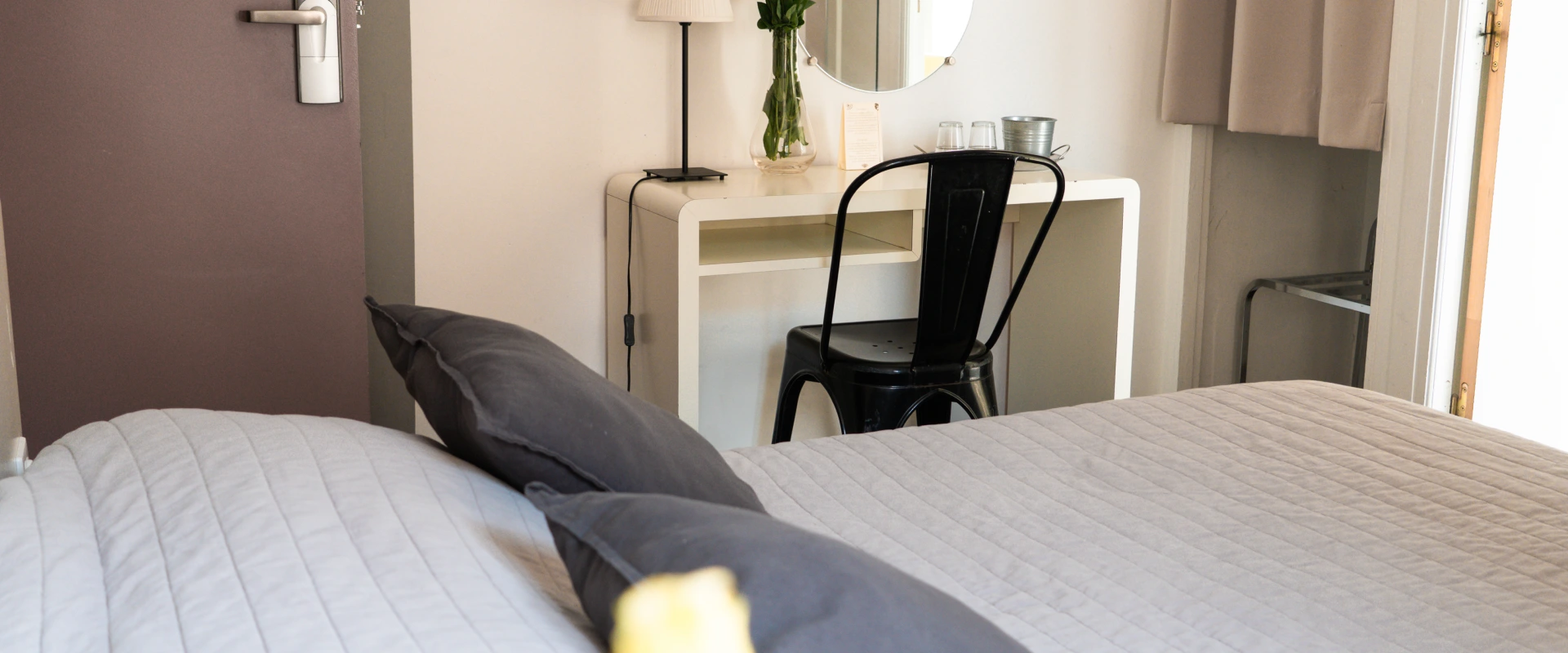 Lit avec couvre-lit gris, bureau blanc, chaise noire, miroir rond, et vase de fleurs.