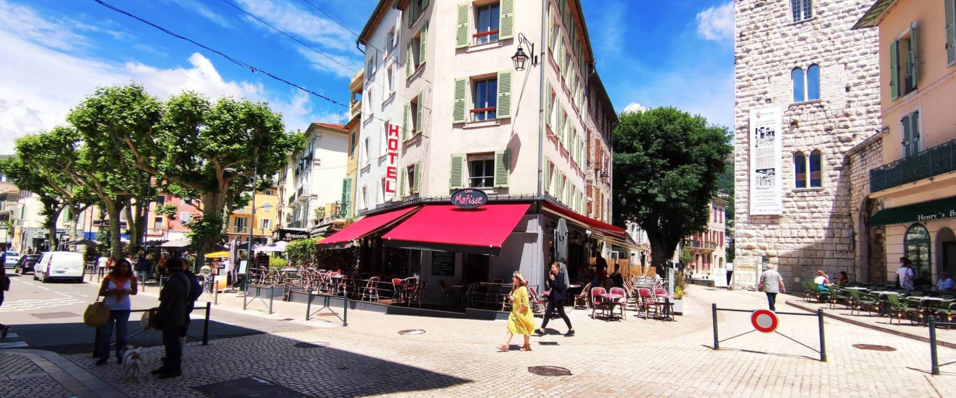 Ruelle ensoleillée avec un café-terrasse et des personnes marchant près d'un bâtiment ancien.