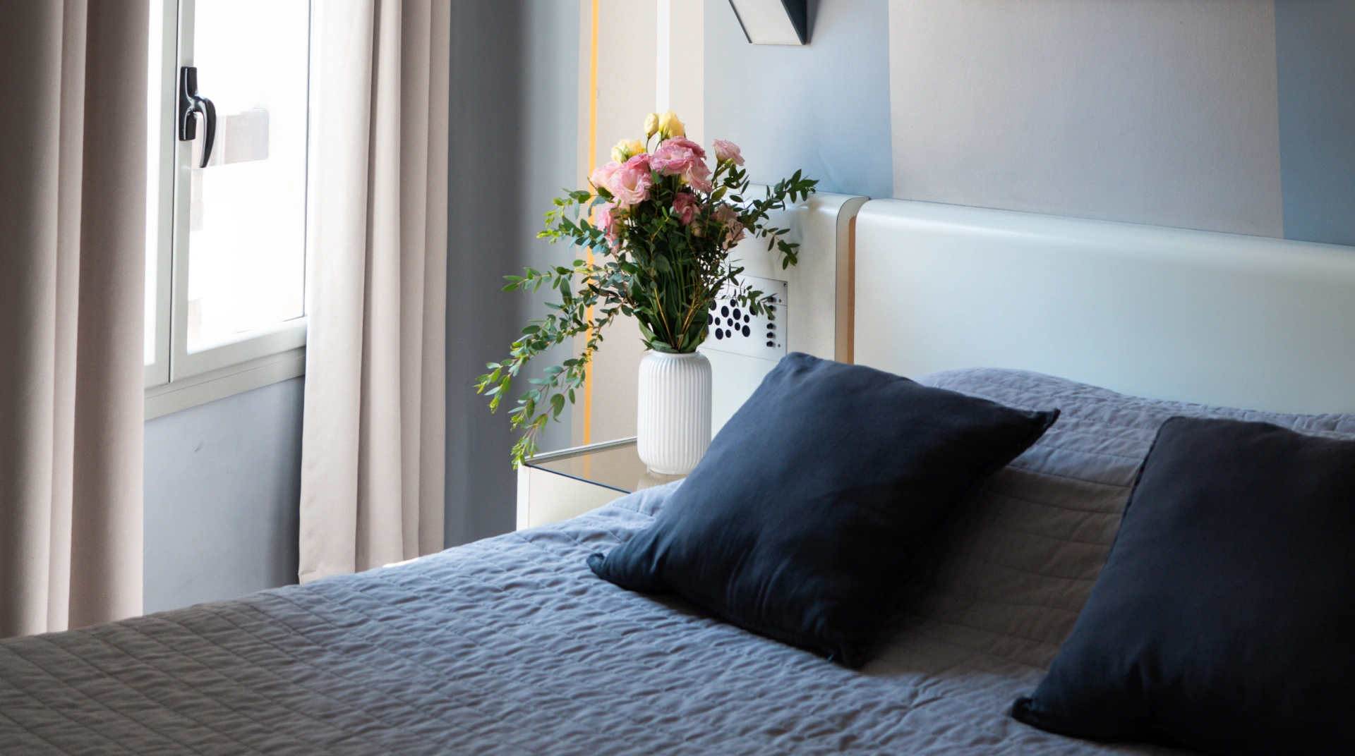 Lit avec couvre-lit gris, coussins foncés, bouquet de fleurs et fenêtre.