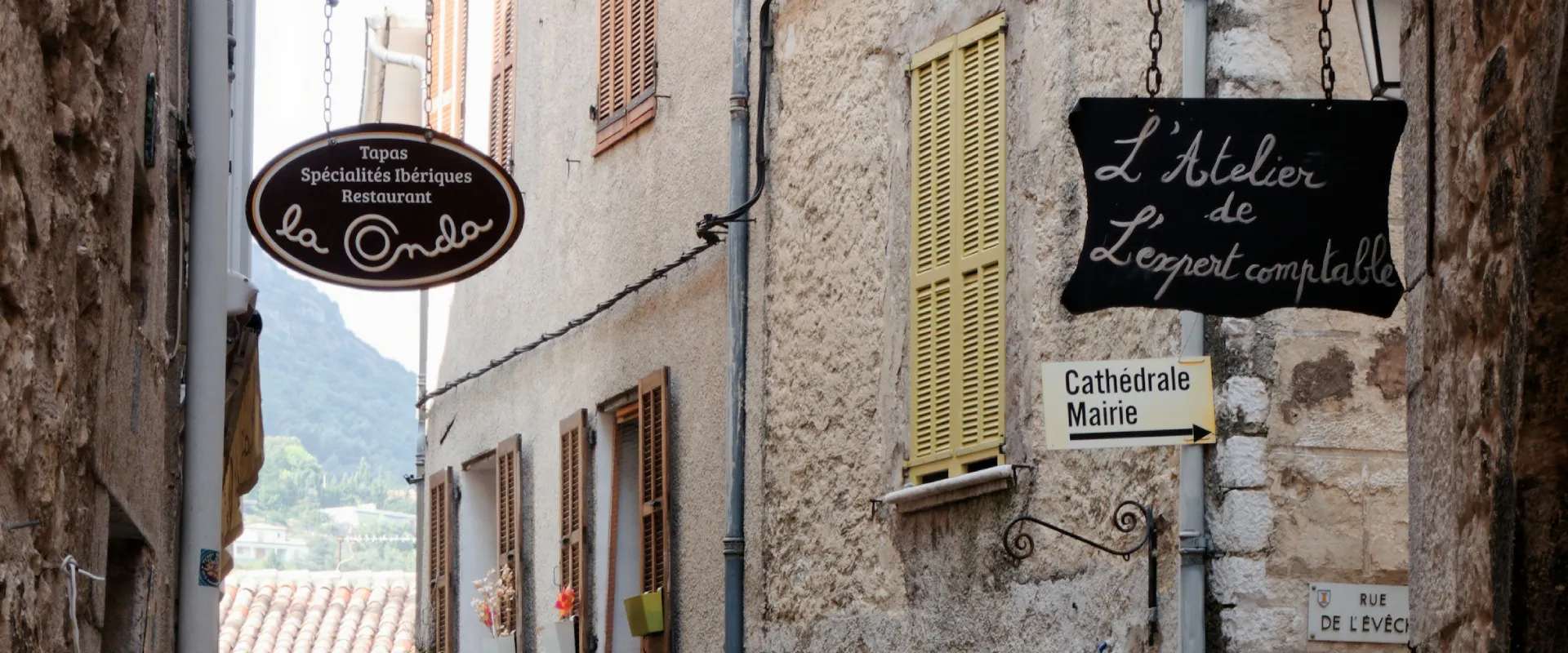 Rue étroite avec enseignes de restaurant et d'atelier, panneau directionnel vers mairie et cathédrale.