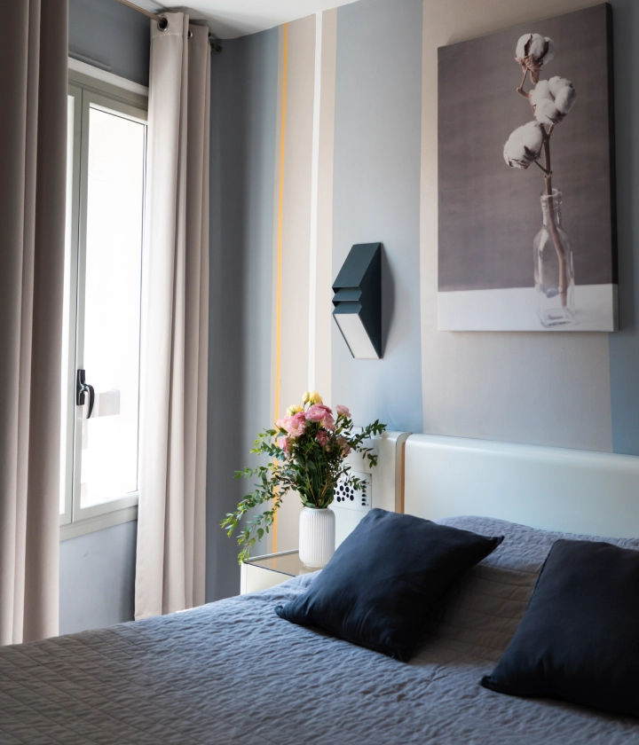 Chambre lumineuse avec lit, coussins bleus, tableau mural et vase de fleurs sur une table de chevet.