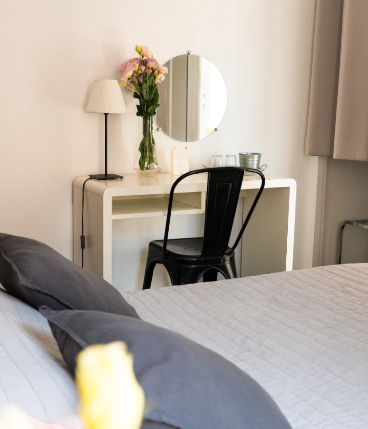Chambre lumineuse avec lit gris, bureau blanc, chaise noire et fleurs dans un vase.