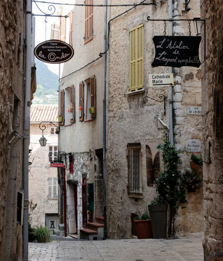 Ruelle pittoresque avec enseignes de restaurants et boutiques, volets colorés, et pavés au sol.