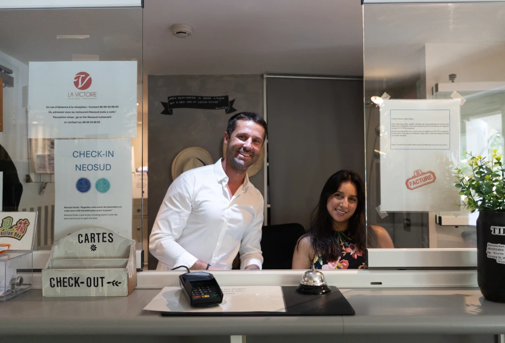 Deux personnes souriantes derrière un comptoir de réception d'un hôtel, avec des affiches visibles.