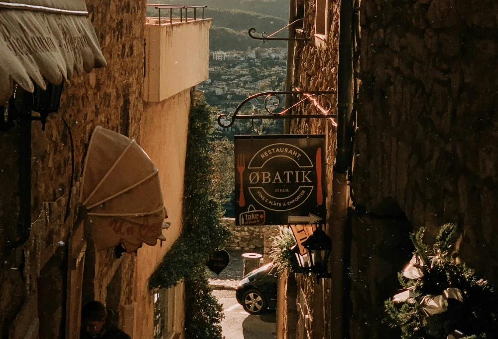 Ruelle en pierre avec panneau de restaurant, ville visible au loin, ambiance ensoleillée.