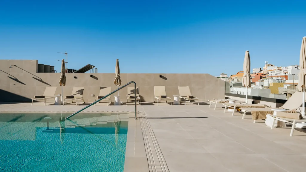 Piscina en azotea con tumbonas, cielo despejado y edificios al fondo.
