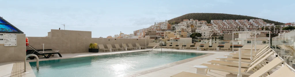 Piscina en la azotea del hotel con tumbonas blancas y vistas al pueblo de Los Cristianos y monta&ntilde;as.