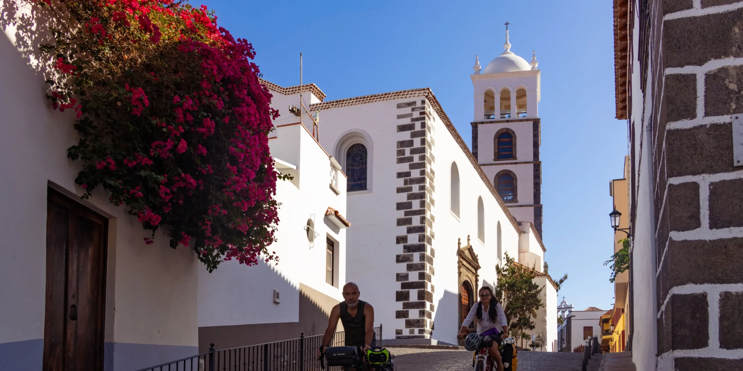 Ciclistas subiendo una calle empedrada en Garachico junto a la iglesia de Santa Ana y fachadas blancas canarias.