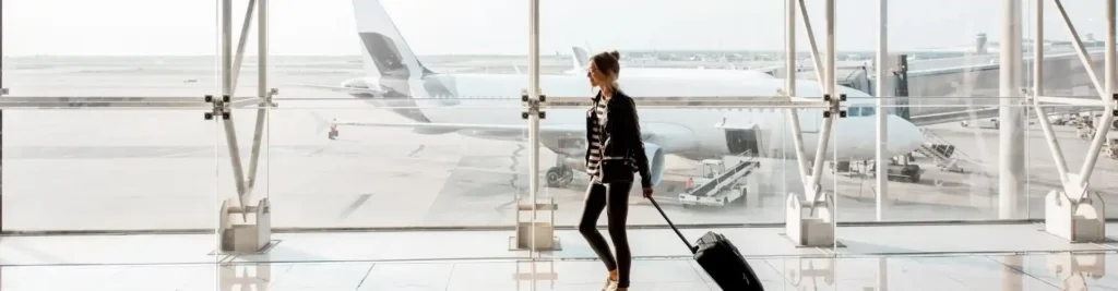 Mujer moderna caminando con maleta de mano en terminal de aeropuerto con avi&oacute;n visible detr&aacute;s.
