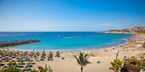 Vista panorámica elevada de la Playa de Los Cristianos en Tenerife. Se observa una bahía de aguas azul turquesa con un barco clásico de velas anclado, una playa de arena dorada con filas de hamacas ordenadas y, a la derecha, un puerto con barcos amarrados. Al fondo se alza un gran acantilado rocoso bajo un cielo despejado.
