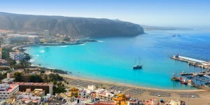 Vista panorámica elevada de la Playa de Los Cristianos en Tenerife. Se observa una bahía de aguas azul turquesa con un barco clásico de velas anclado, una playa de arena dorada con filas de hamacas ordenadas y, a la derecha, un puerto con barcos amarrados. Al fondo se alza un gran acantilado rocoso bajo un cielo despejado.