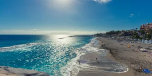 Vista a nivel de la Playa del Duque en Tenerife bajo un sol radiante que se refleja intensamente en el océano Atlántico. El agua muestra tonos azules intensos y espuma blanca al romper en la orilla de arena oscura. Personas caminan y nadan a lo largo de la costa, mientras que la orilla cuenta con áreas de sombrillas y hamacas cerca de complejos hoteleros.