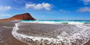 Paisaje natural de la Playa de La Tejita en Tenerife. En primer plano, las olas del mar rompen dejando un semicírculo de espuma blanca sobre la arena oscura y húmeda. Al fondo, a la izquierda, destaca la silueta rojiza de la Montaña Roja bajo un cielo azul brillante con nubes blancas dispersas.
