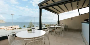 Terraza con mesas y sillas blancas bajo una pérgola, con vistas panorámicas al mar y al puerto.