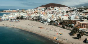 Aerial view of a wide urban beach in Los Cristianos, Tenerife, full of bathers, umbrellas, and sun loungers arranged on the sand. In the calm water, some people can be seen swimming and kayaks near the shore. In the background is a dense urban core of white buildings ascending the slope of a volcanic hill.