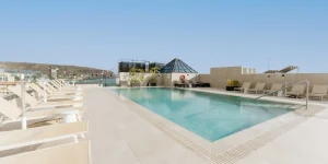 Rooftop con piscina del hotel Reverón Plaza con una piscina rectangular de agua cristalina en el centro. A ambos lados de la piscina hay alineadas hamacas de color beige sobre un suelo de baldosas claras. Al fondo se aprecia una estructura piramidal de cristal, vegetación en maceteros y vistas lejanas a las montañas costeras bajo un cielo despejado.