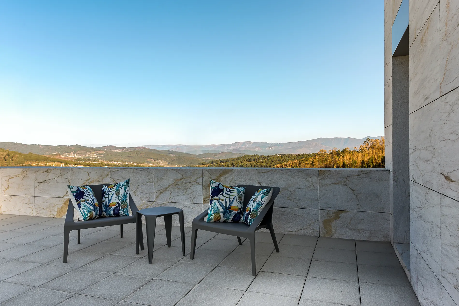 Terra&ccedil;o com cadeiras, mesa e vista panor&acirc;mica para colinas ao fundo.