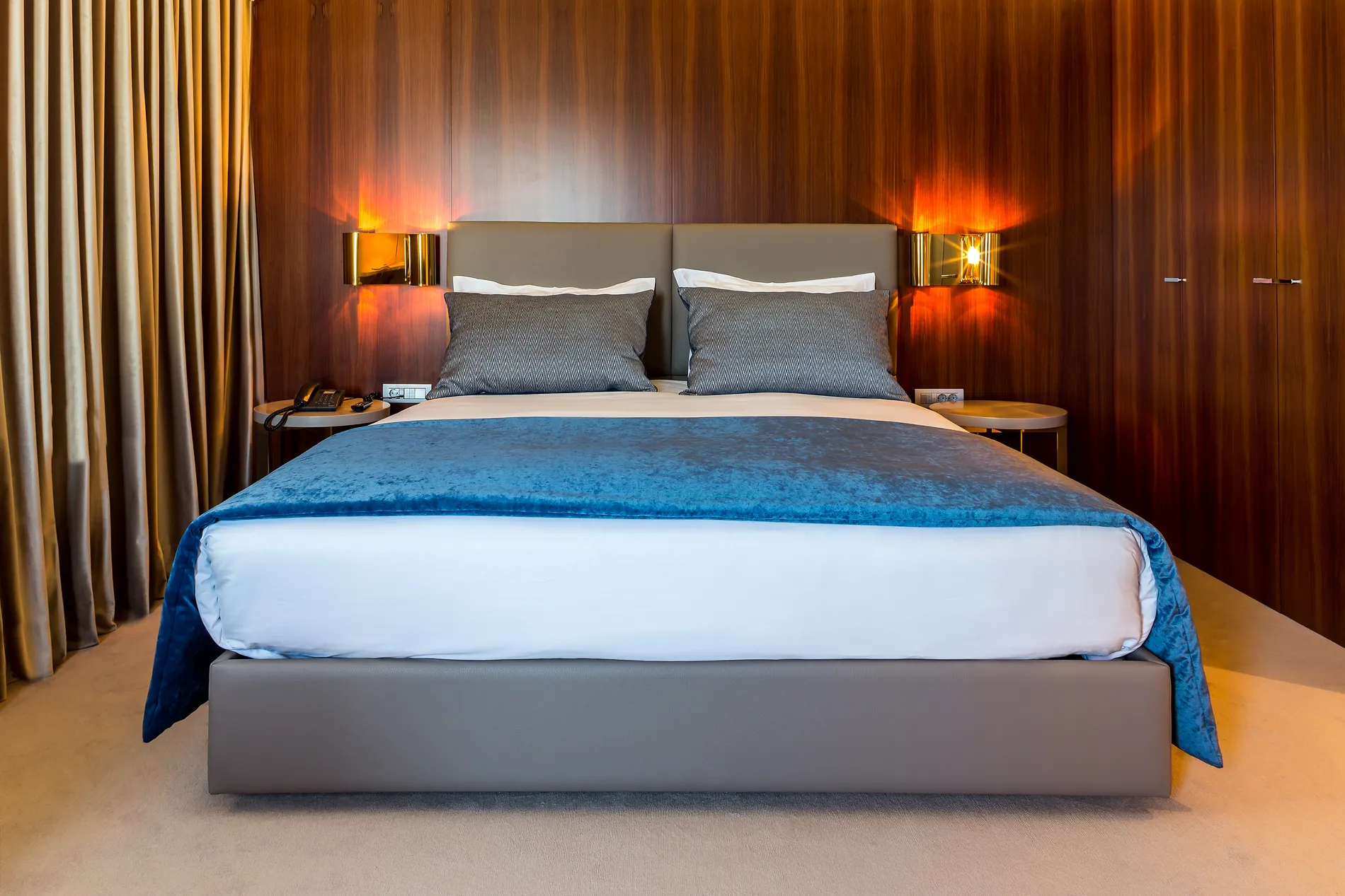 Cama arrumada com cobertor azul e lumin&aacute;rias acesas na cabeceira.