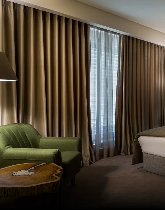 Quarto de hotel com cadeira verde, mesa de madeira e cama ao lado da janela.